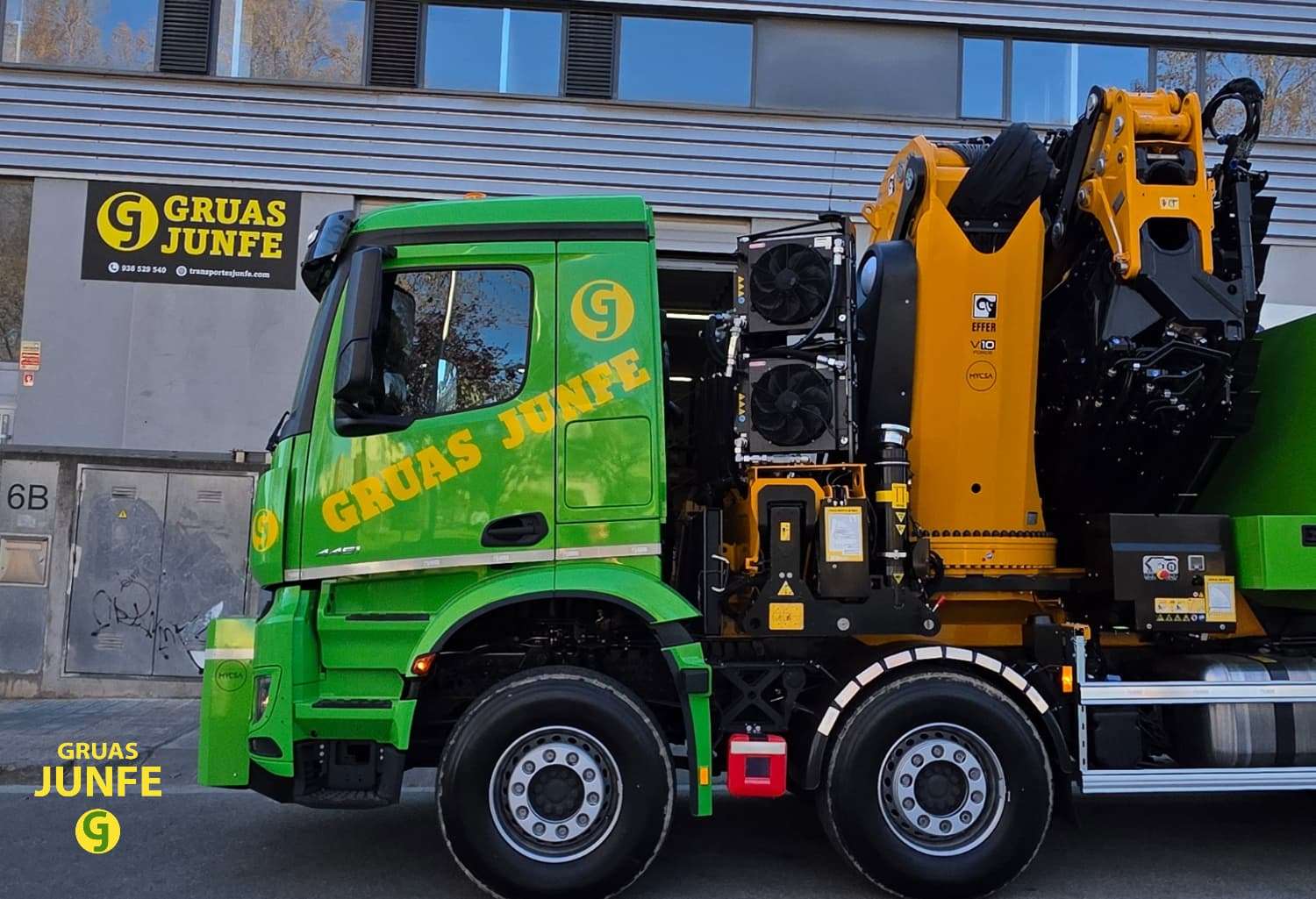 Vista lateral de un moderno camión grúa articulado de alta capacidad de la flota de Grúas Junfe en Barcelona, aparcado frente a sus instalaciones.