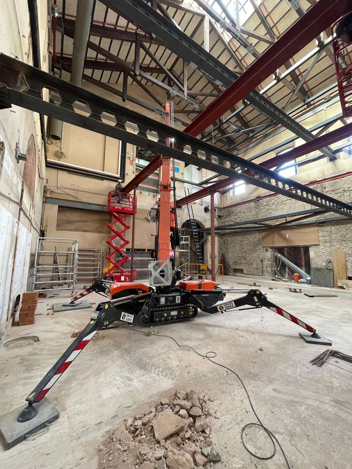 Detalle de la estabilización y maniobra de una mini grúa naranja en el interior de una estación en obras.