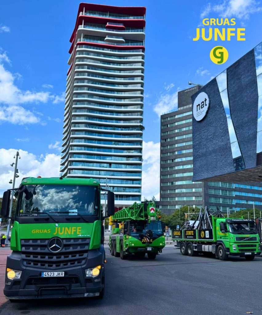 Flota coordinada de Grúas Junfe con grúa móvil de celosía y camiones de transporte en gran avenida con rascacielos.