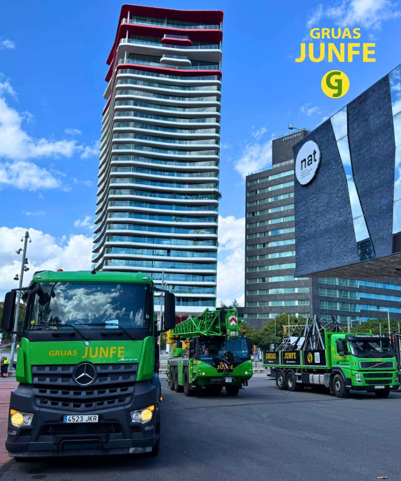 Flota coordinada de Grúas Junfe con grúa móvil de celosía y camiones de transporte en gran avenida con rascacielos.